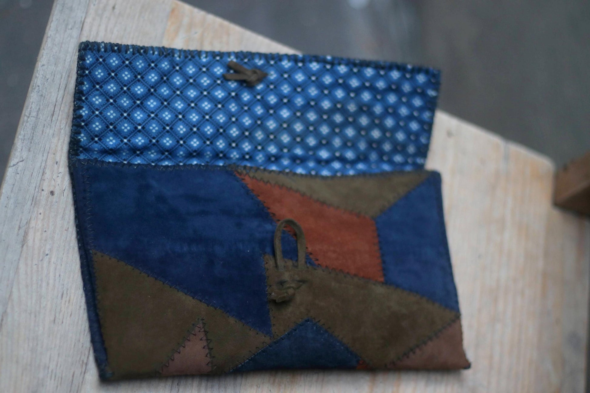 A vintage clutch made of patchwork suede in various earthy tones, including navy, brown, green, and terracotta. The clutch has a wooden toggle closure and is resting on a light-coloured surface. the inside lining is blue
