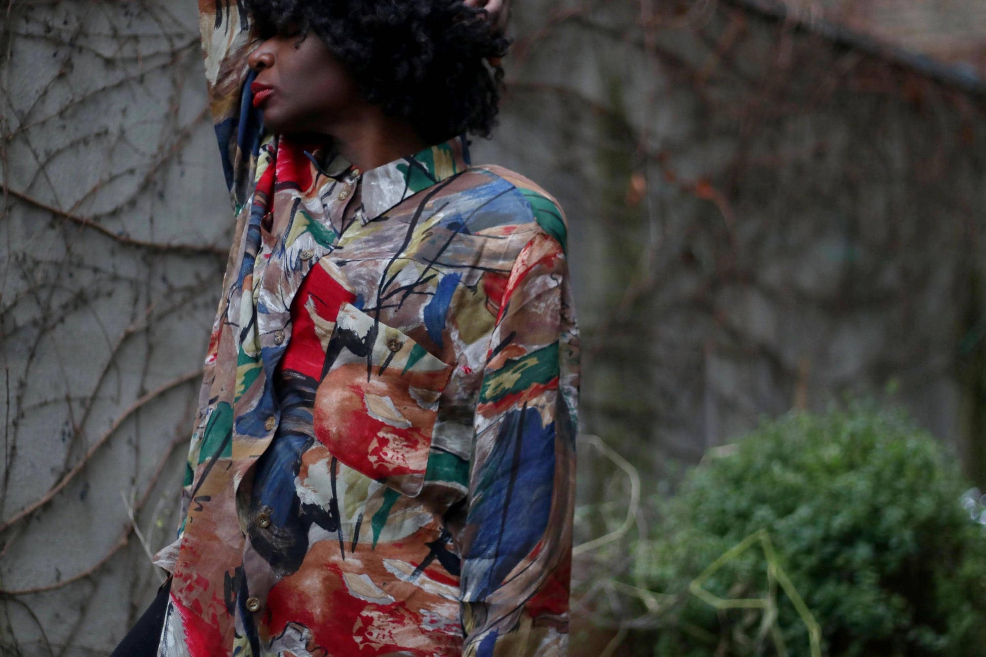A stylish individual poses against a textured concrete wall entwined with bare vines, wearing a vintage Gerry Weber oversized button-up blouse featuring a bold abstract print in red, blue, green, and neutral tones.