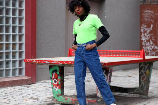 Woman in high-waisted vintage blue jeans with a relaxed straight-leg fit, paired with a neon green tee under a black long-sleeved turtleneck is photographed against an urban background, including a graffiti-covered ping pong table