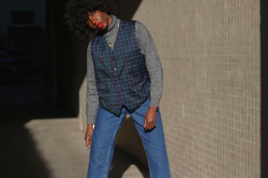 A stylish individual wears a vintage blue checkered vest over a gray turtleneck, paired with wide-leg denim. The vest features a structured fit, deep navy plaid pattern, and fine green lines. The subject leans casually against a sunlit wall.