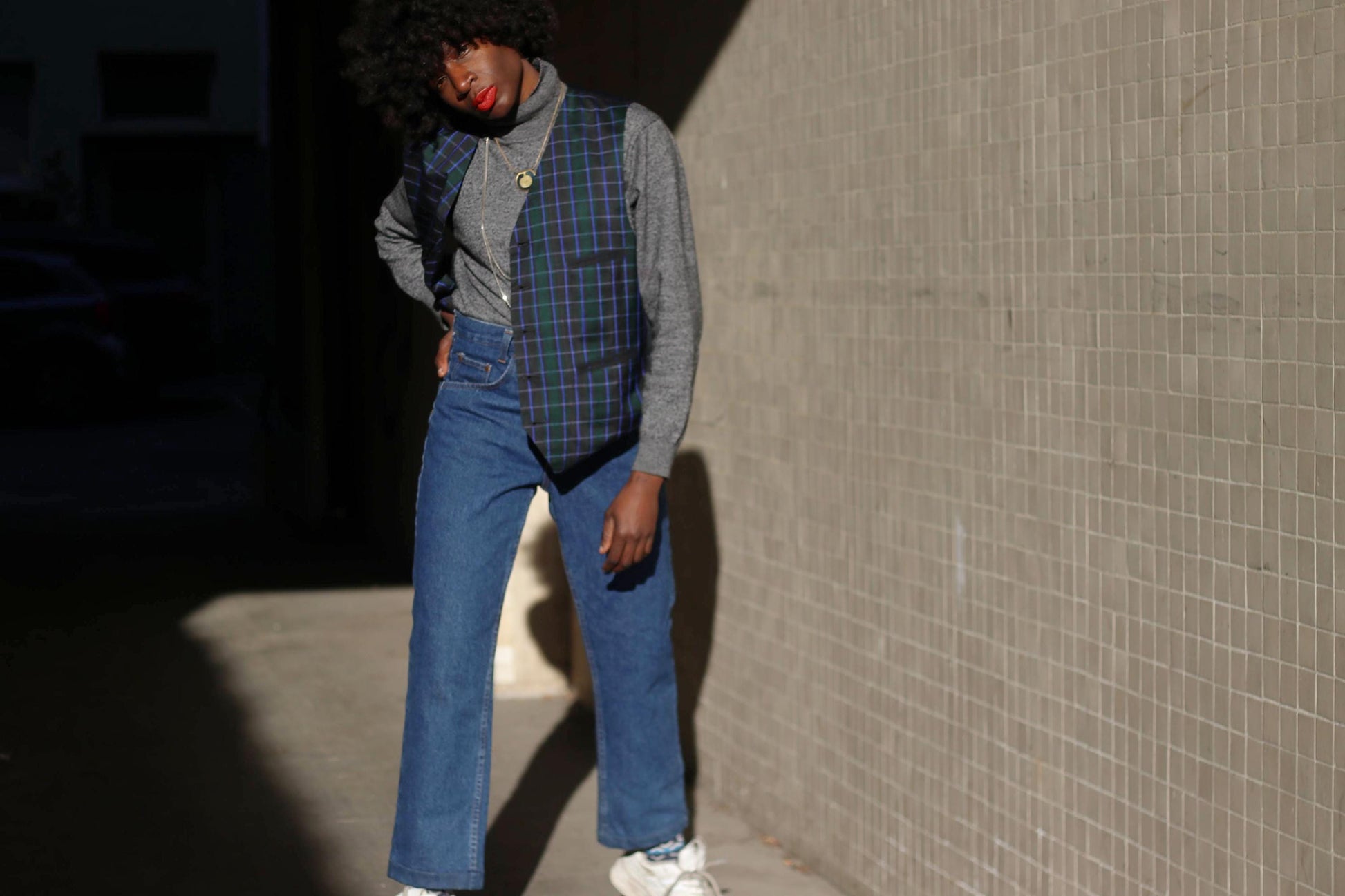 A stylish individual in a vintage plaid vest in navy and green  layered over a fitted sweater, embodying a mix of preppy and grunge fashion against a modern architectural backdrop.