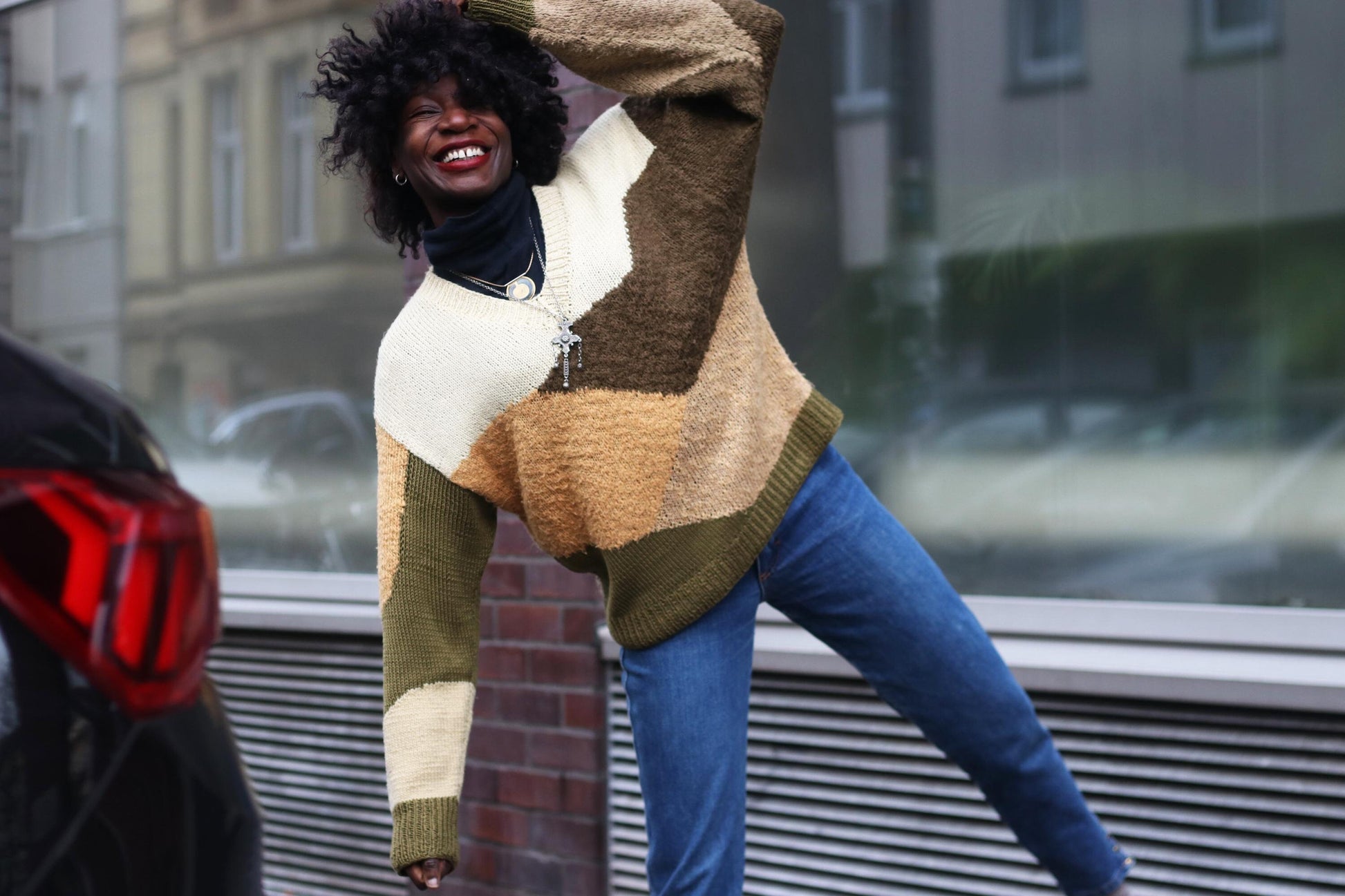 Smiling female model wearing a colorful  vintage handmade patchwork sweater  in brown and green with blue jeans, playfully posing on a city street.
