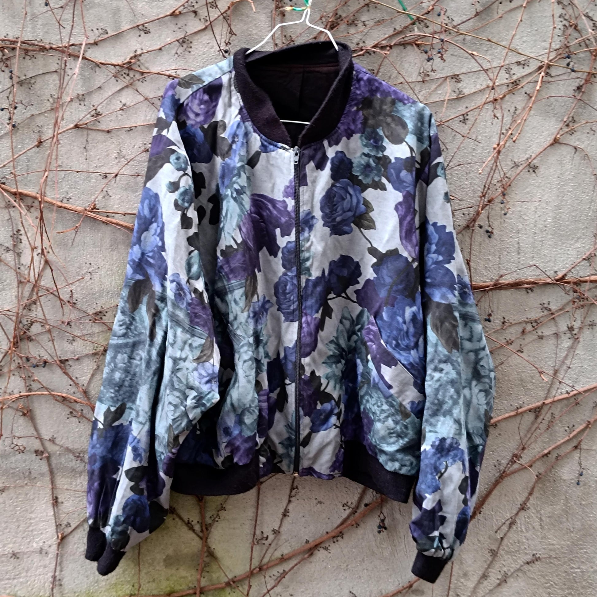Vintage y2k bomber jacket with a blue and purple watercolor floral print. Relaxed fit, dark ribbed cuffs and hem, silver zipper. Hung against a textured wall with vines