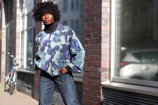 Female model wearing retro blue jeans, black turtleneck and a vintage bomber jacket with a blue and purple watercolor floral print. She is photographed outdoors in front of a large window and near a brick wall.