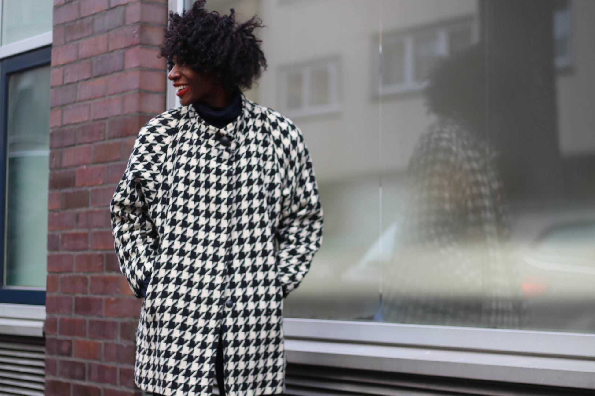A woman with curly black hair earing a vintage black-and-white houndstooth cashmere-wool coat, standing against a city backdrop