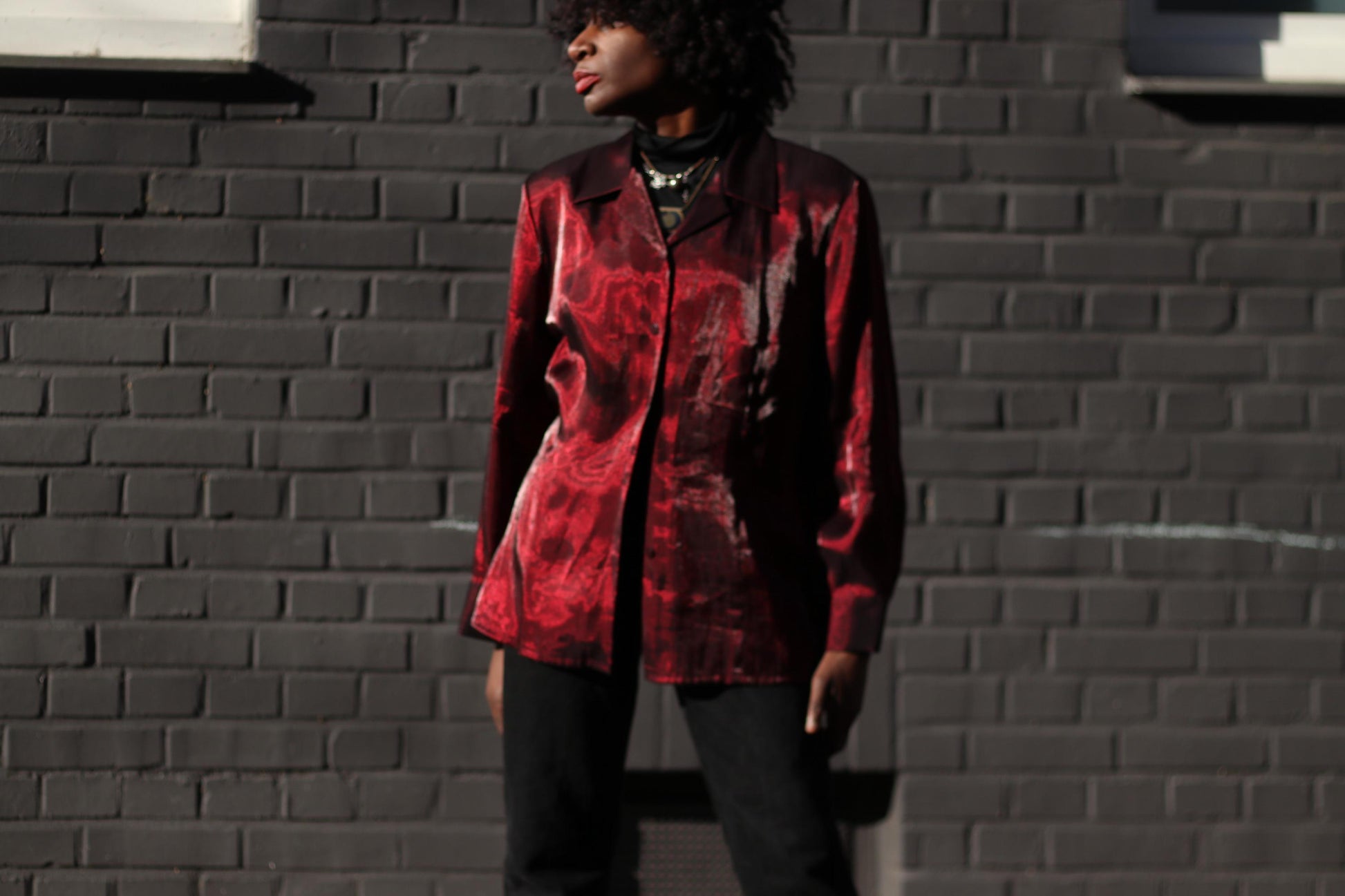 Female Model of African heritage in  a glossy red Y2K vintage blouse styled with black jeans and turtleneck captured against a dark brick wall in direct sunlight.