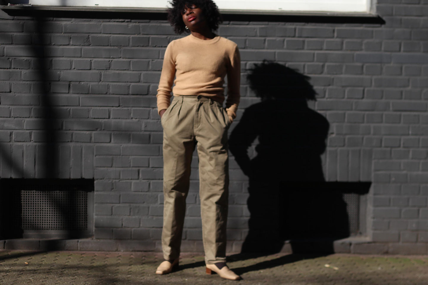 A female model wearing vintage high-waisted khaki cargo pants, styled with a fitted beige sweater, standing against a modern industrial brick wall