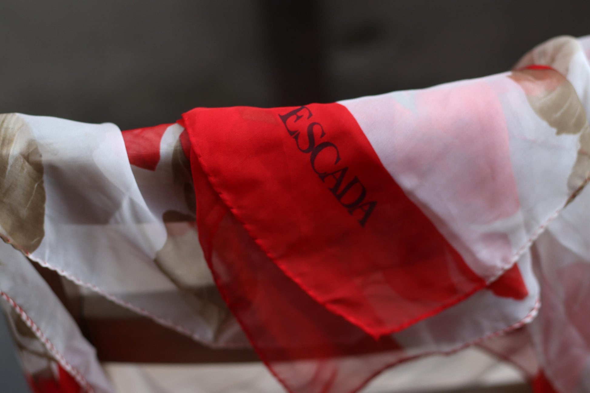 Close-up of the ESCADA logo on a Soft ivory silk scarf with vibrant red florals, representing classic 1980s Escada style, elegantly draped on a wooden cha