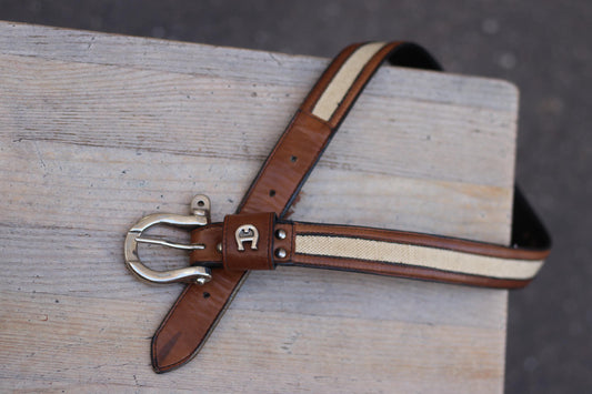 A vintage Etienne Aigner belt laid flat against a white background, showcasing its brown leather with a cream stripe and a gold-tone buckle.
