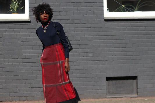A stylish woman wearing a red plaid maxi skirt and black fitted top, standing against a modern gray brick wall, exuding effortless elegance.