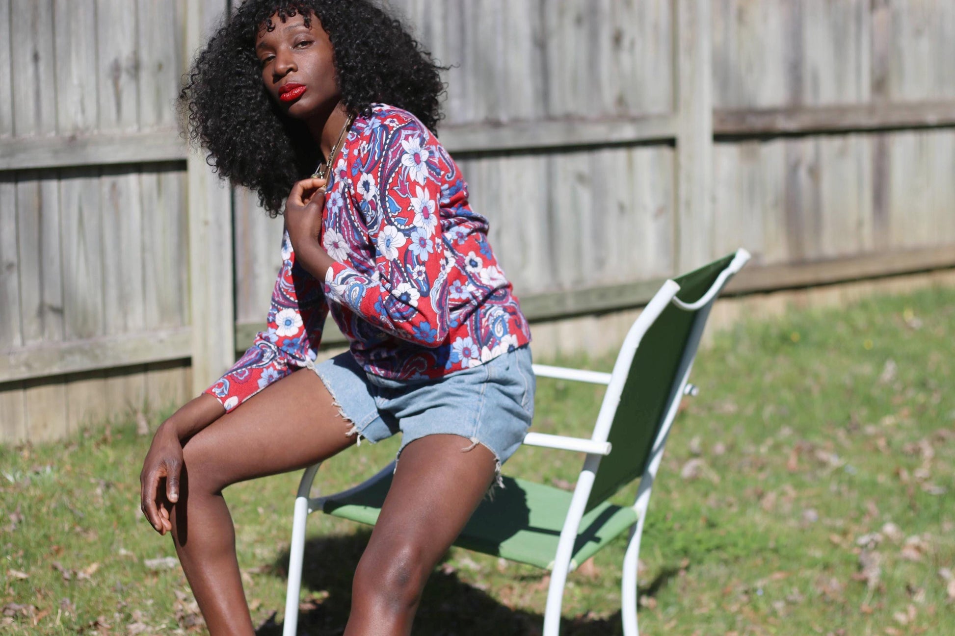 A female model with curly black hair sitting on a white and green lawn chair on a sunny lawn, showcasing a red, white, and blue patterned vintage Talbot's blouse made of 100% merino wool. A wooden fence and green foliage form the backdrop.