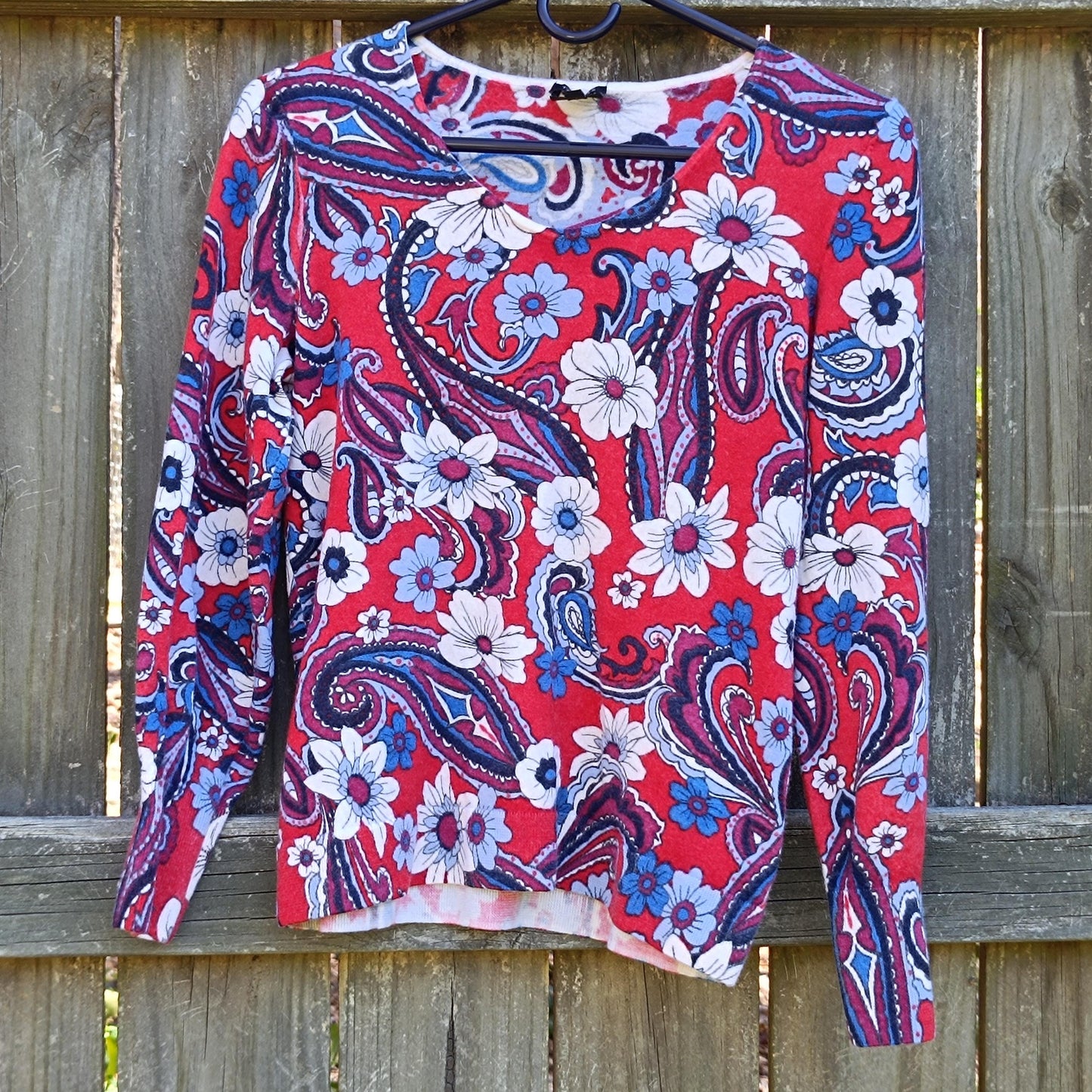 The back view of a vintage merino wool Talbot's blouse. The red, white, and blue long-sleeved floral top is photographed on a hanger against a brown wooden fence outdoors