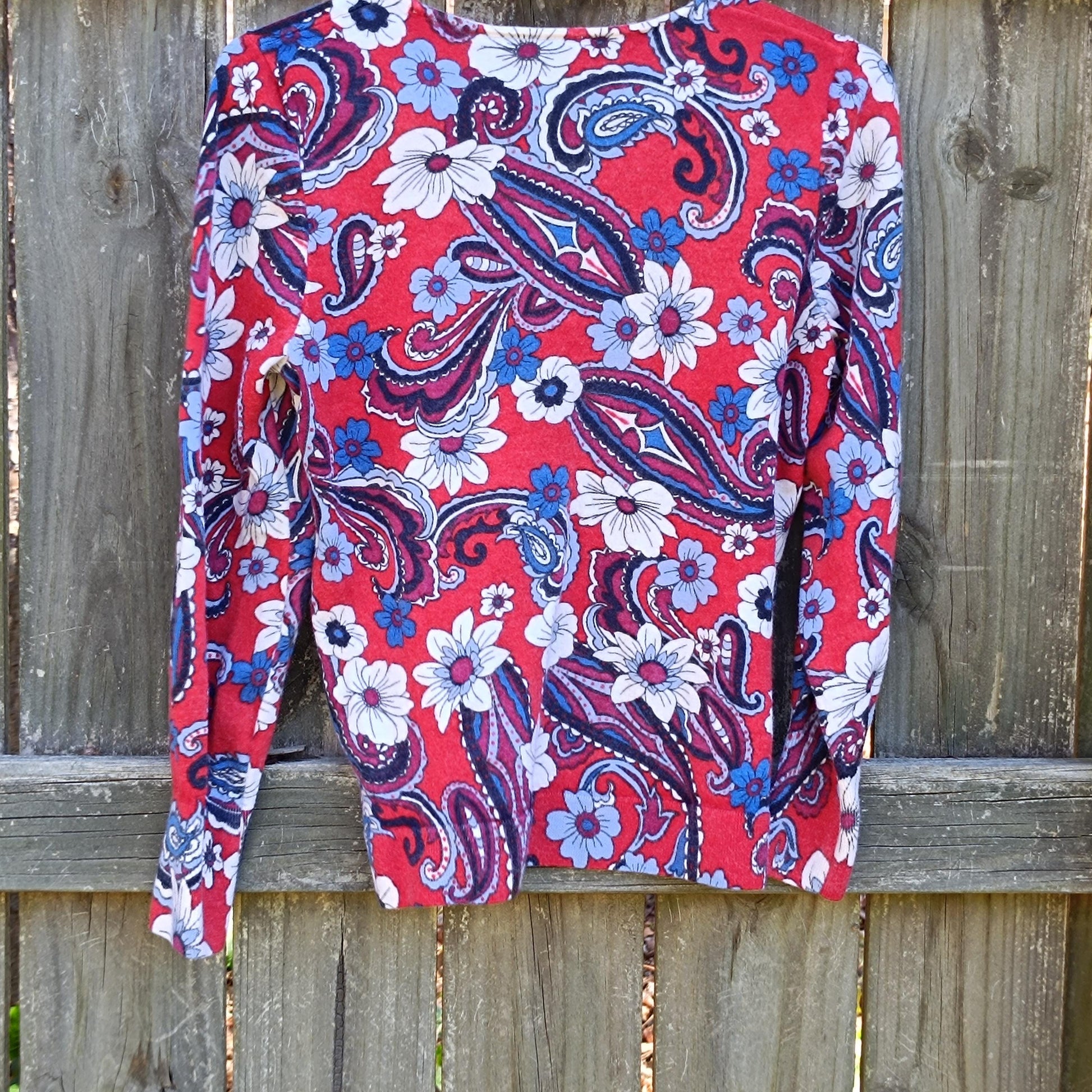 Vintage merino wool Talbot's blouse with red, white, and blue floral patterns is photographed on a hanger  against a brown wooden fence outdoors
