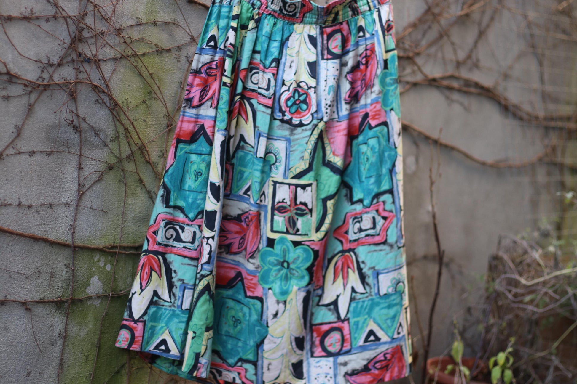 Vintage 80s skirt with a bold, colorful abstract floral print, showcased on a hanger against a textured stone wall with climbing greenery.