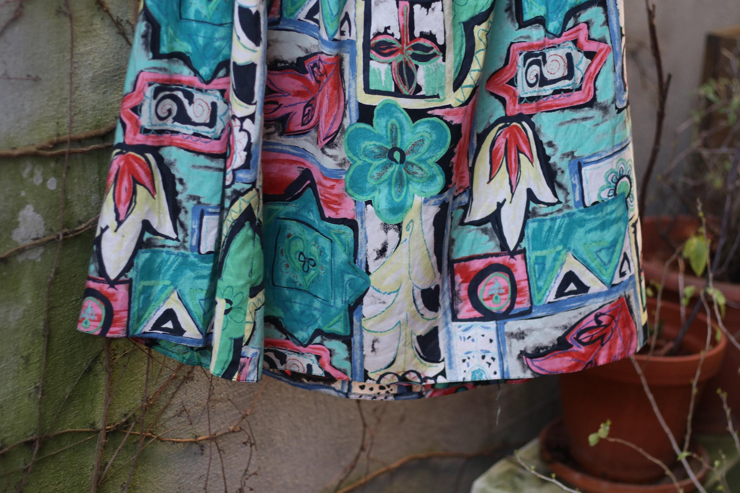 A brightly patterned 80s vintage midi skirt is displayed on a hanger against a rustic stone wall with overgrown vines