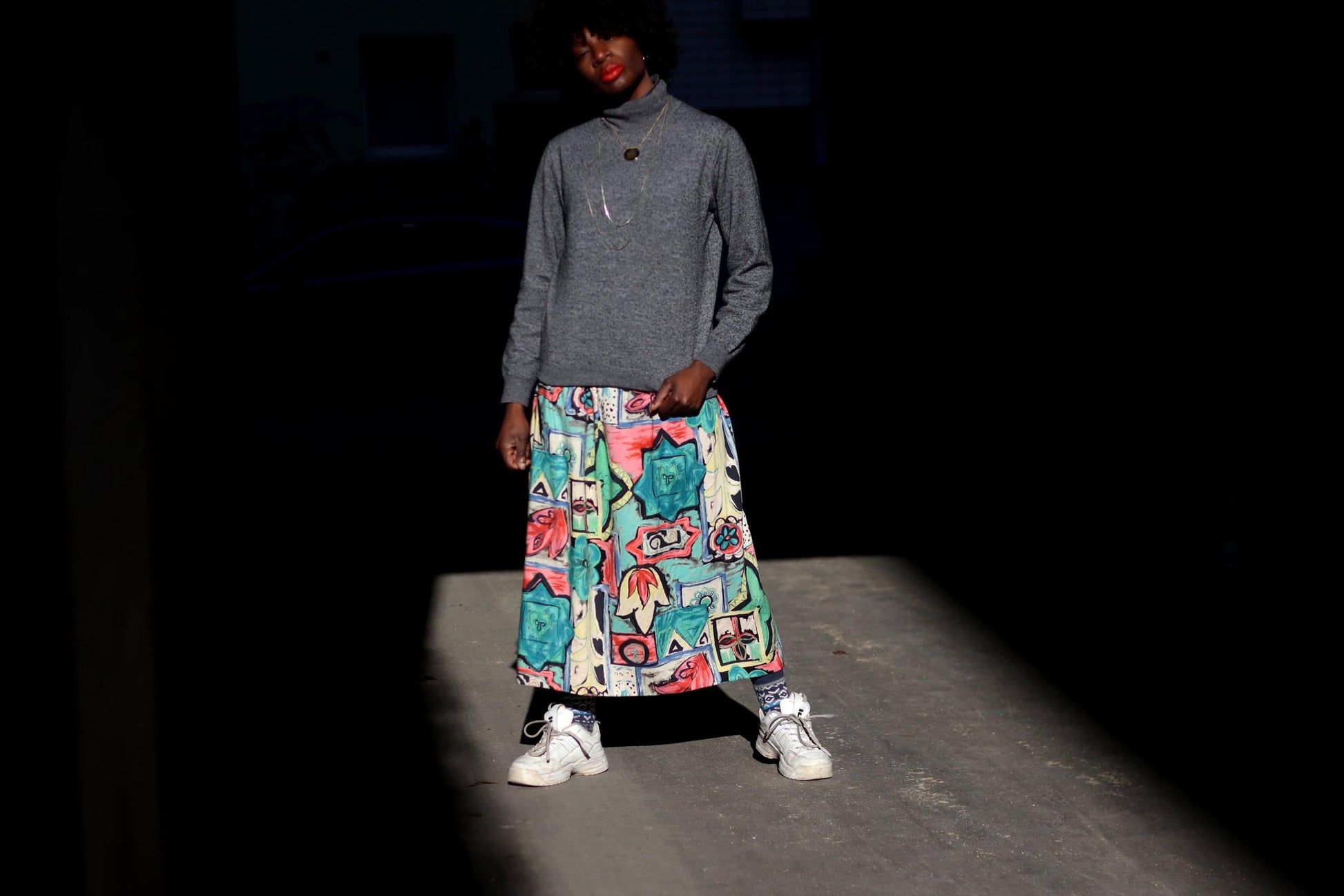 A female model with curly black hair wears a long, vintage skirt with a unique, artistic print against a dark, shadowy background. They are dressed in a gray sweater and white sneakers