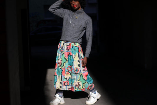 A female model stands against a dark backdrop wearing a long, vintage skirt with a colorful, abstract pattern. They are also wearing a gray turtleneck sweater and white sneakers