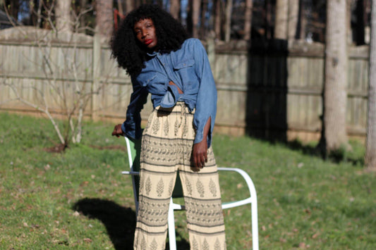 Female model with curly black hair in  a vintage hippie-chic lounge pants and denim shirt photographed outside in a garden next to a green lounge chair. The vintage outfit is  perfect for a relaxed yet statement-making outfit.