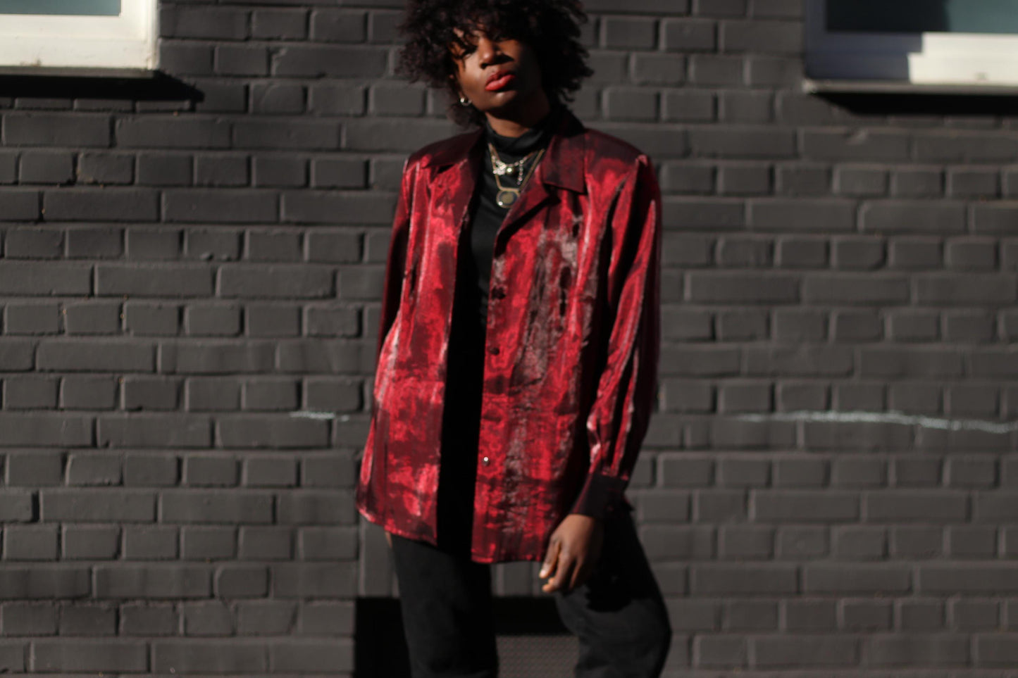 Female model of African heritage in a shiny red button-up blouse by the vintage brand TRU. The shirt has  a strong collar and shoulder pads and is styled with layered necklaces for a bold Y2K look.