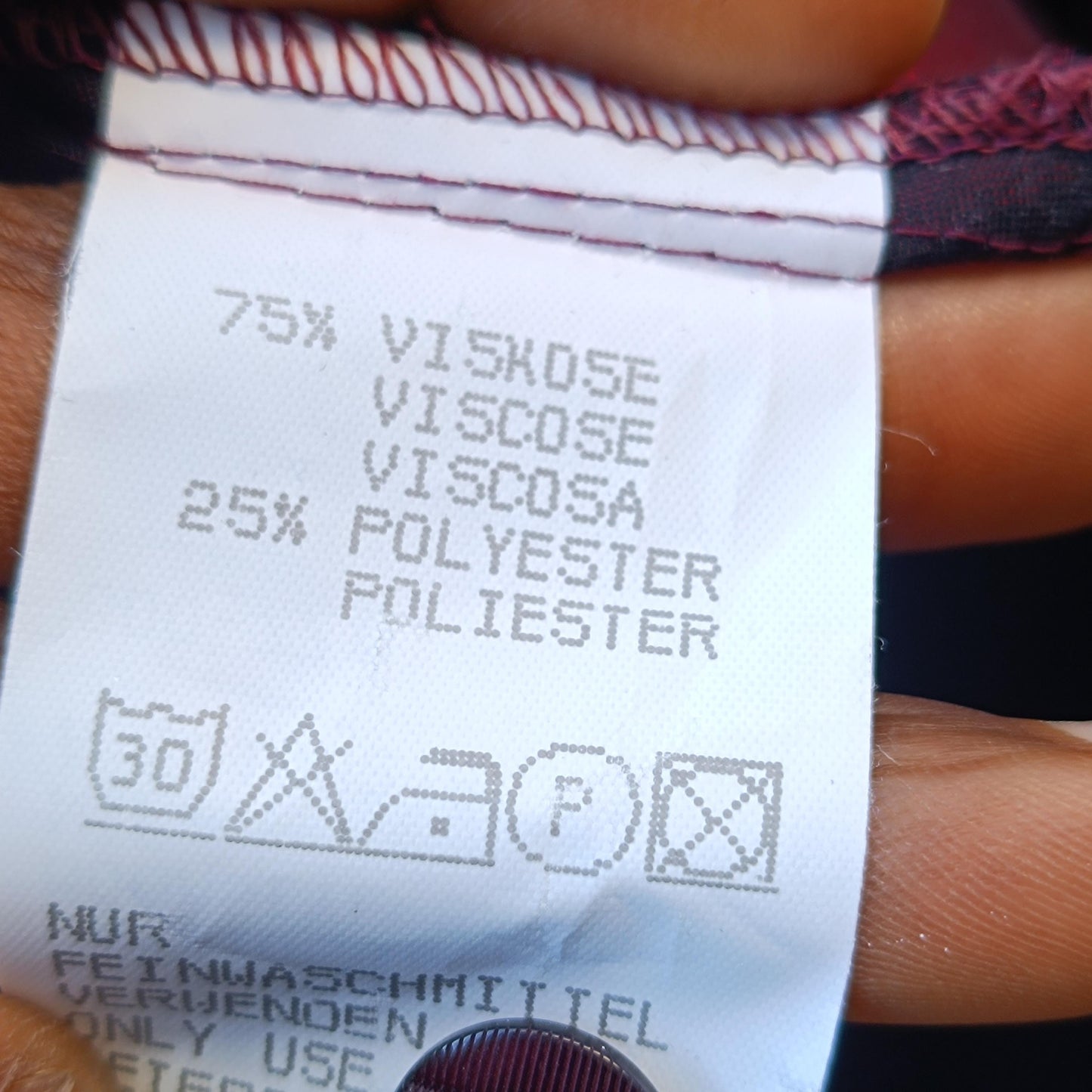 closeup of the fabric tag of the glossy red Y2K vintage blouse. Tag reads 75% viscose 25% polyester. Care instructions to wash at 30 degrees , no drying and ironing allowed is also displayed
