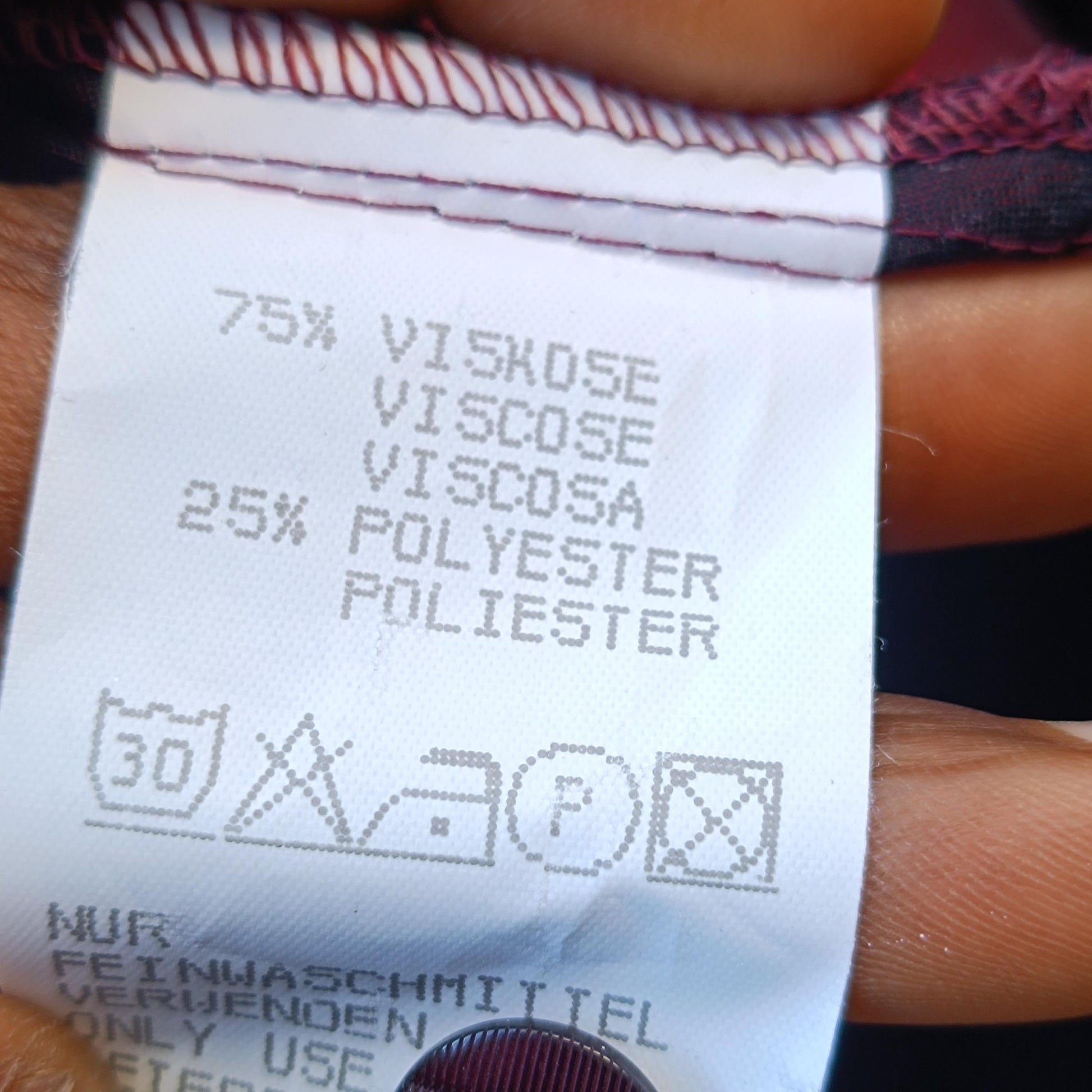 closeup of the fabric tag of the glossy red Y2K vintage blouse. Tag reads 75% viscose 25% polyester. Care instructions to wash at 30 degrees , no drying and ironing allowed is also displayed