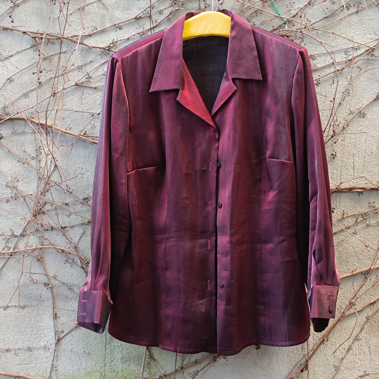 Vinesge glossy red Y2K blouse photographed hanging from a yellow hanger against a light brick wall covered in brown twines