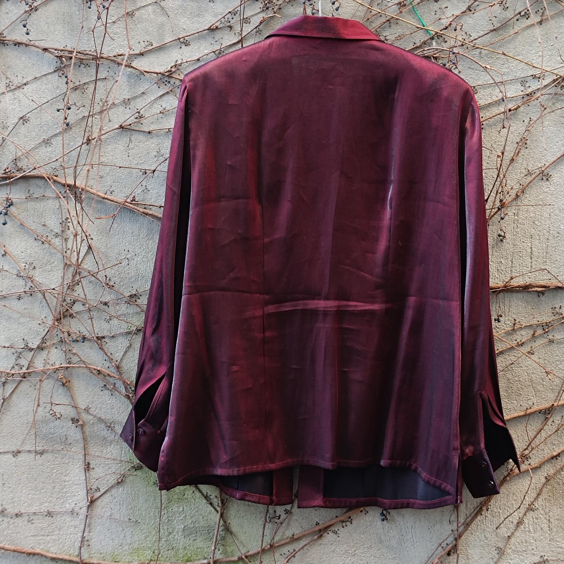 Back view of a vintage Shiny red button-up blouse with strong collar and long sleeves captures hanging from a clothing line outdoors against an ivy covered brick wall.