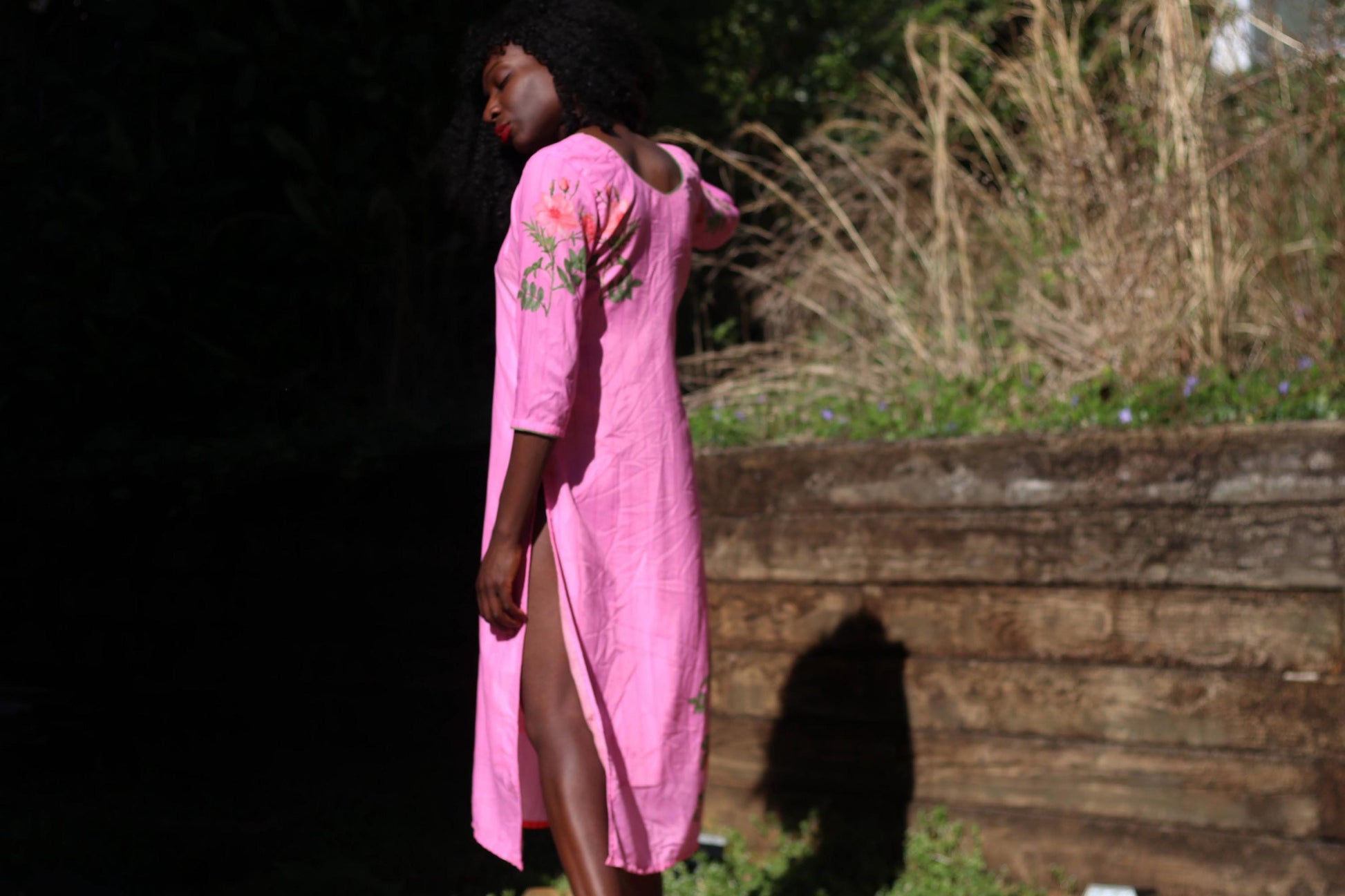 Women wearing an elegant pink vintage dress with floral embroidery and Southeast Asian craftsmanship photographed against a country-inspired background with browning grass and wooden structures