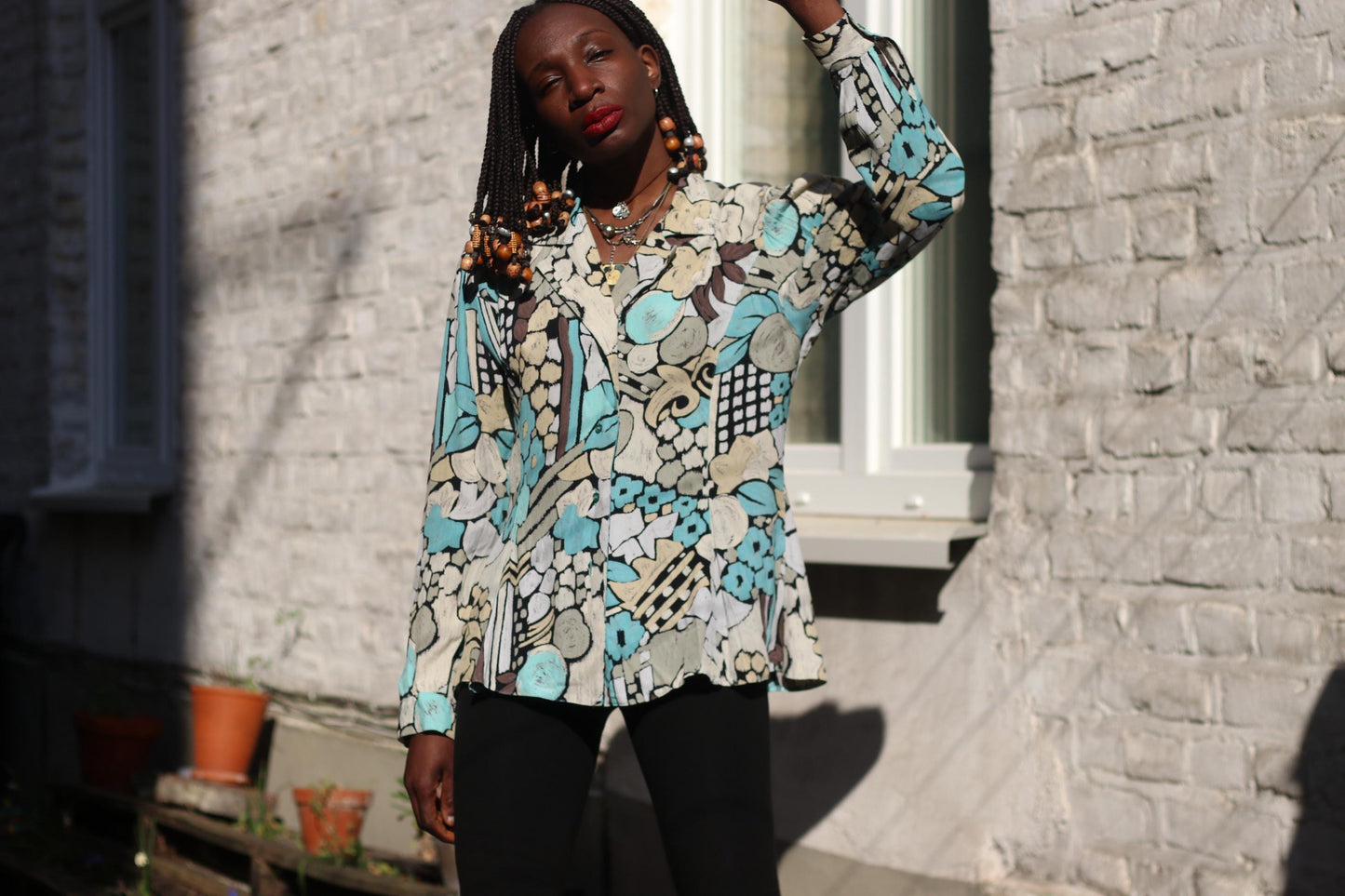 A model with dark skin and braided hair adorned with wooden beads poses outdoors in sunlight, wearing a vintage viscose blouse with an abstract print in blue, beige, and black, against a textured brick wall and a window.