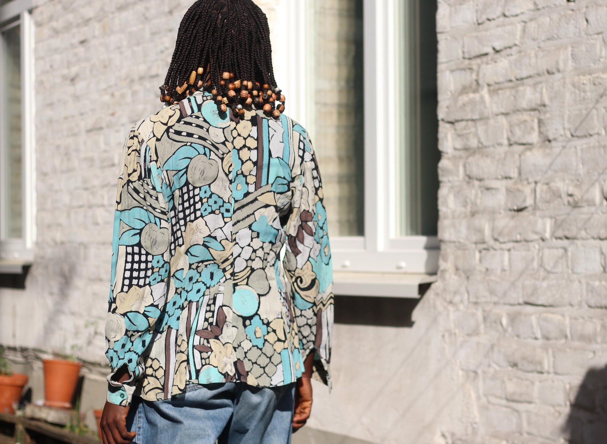 The back of a stylish in the afternoon sun, showcasing a fluid, long-sleeved 90s blouse with an artsy geometric pattern, her braided hair adorned with wooden beads.
