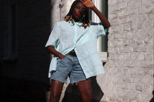 A female models a light blue linen blouse and denim shorts outdoors, posing with one hand raised near their face. The blouse is loose-fitting and the shorts are above knee-length.