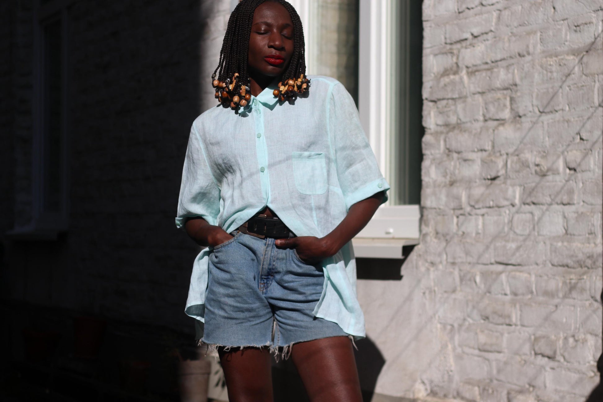 A female model of African heritage stands outside wearing a light blue, relaxed-fit, vintage linen blouse and denim shorts. The blouse is airy and the shorts are mid-thigh length
