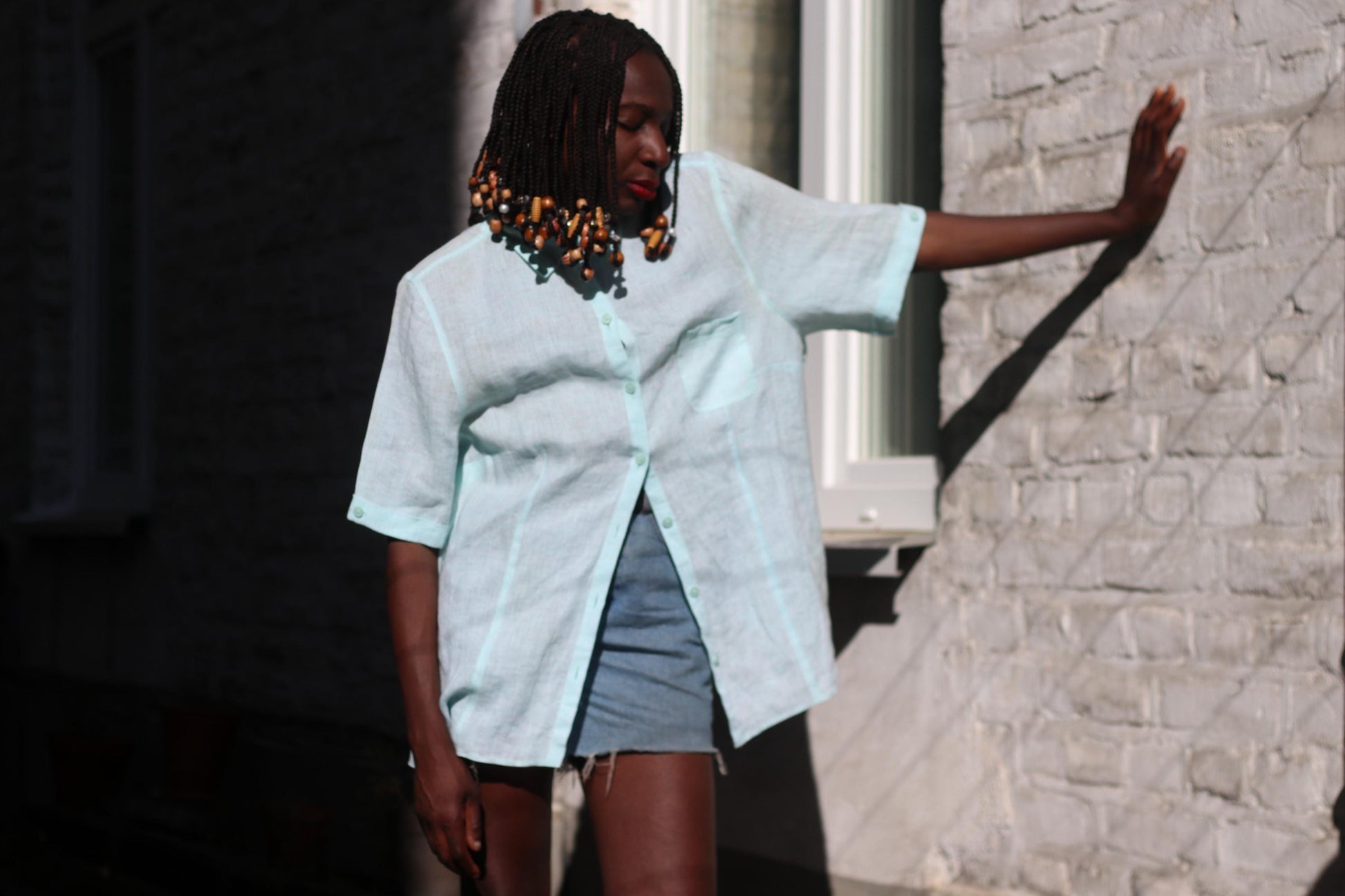 A female model of African heritage showcases a light blue vintage linen blouse and denim shorts outdoors, with a brick wall and sunlight in the background. The vintage 90s blouse is short-sleeved and the shorts are casual.