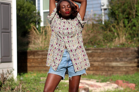 A female model wearing a patterned Vintage Pierre Cardin short-sleeved shirt and denim shorts is photographed outdoors. The shirt has a geometric design in shades of beige, red, and gray. The person&#39;s arms are raised above their head.
