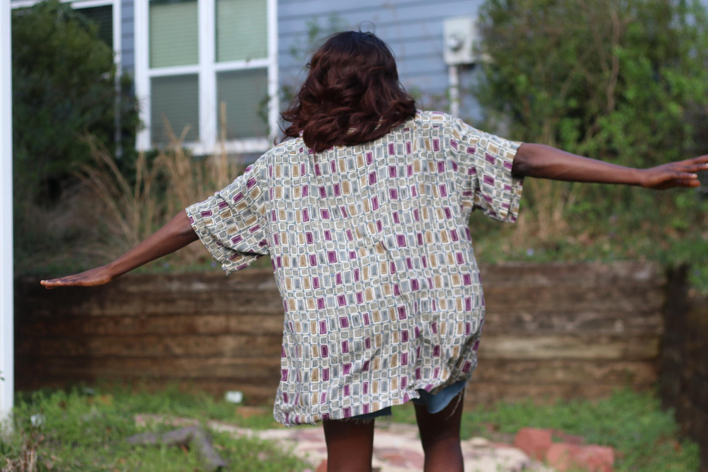 A female model in a button-down  Pierre Cardin shirt with an abstract pattern and light blue denim shorts is captured from behind  outdoors.