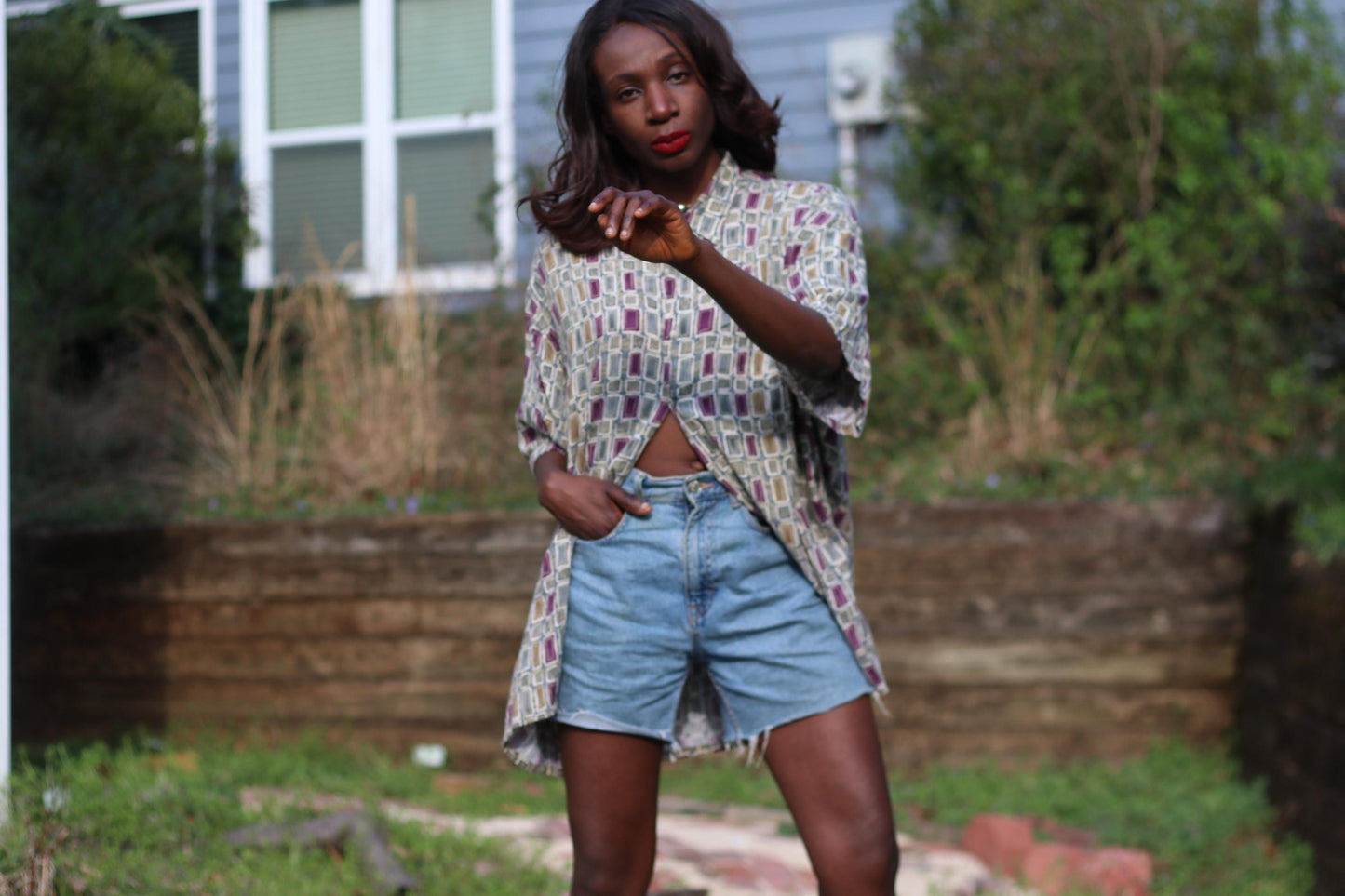 A model of African heritage  wearing a short-sleeved shirt vintage Pierre Cardin shirt from the 90s with a geometric print and denim shorts is photographed in an outdoor setting. The shirt is beige with red and gray rectangles. The person&#39;s left arm is slightly raised, and they have dark hair.