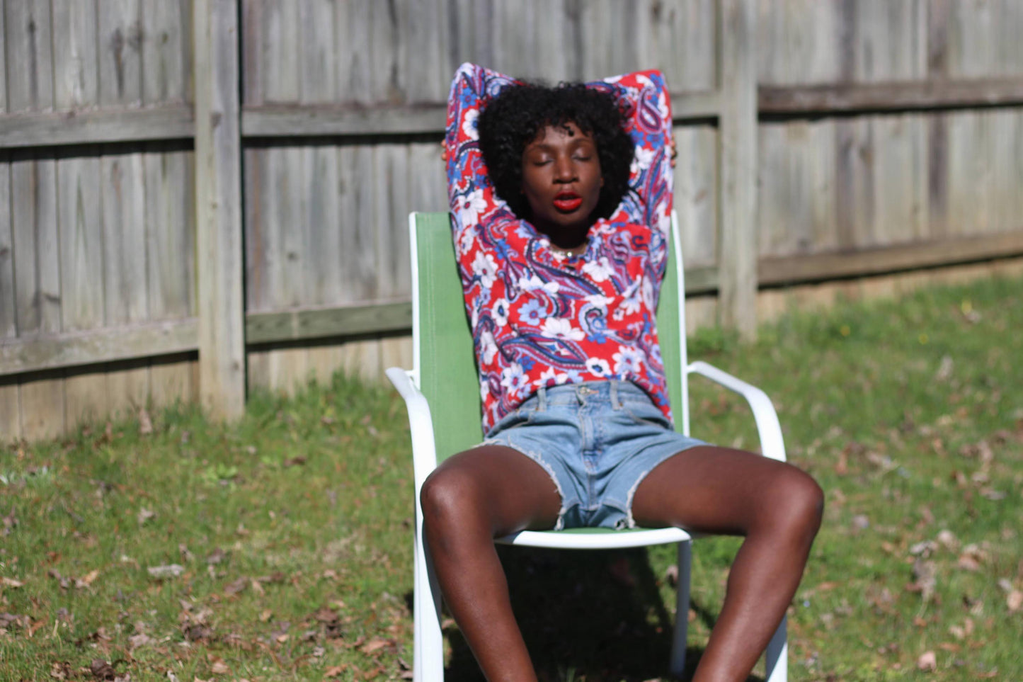 In a bright outdoor setting with a green lawn and a wooden fence, a woman is seated, displaying a vintage long-sleeved blouse by Talbot&#39;s. The retro  blouse features a colorful red, white, and blue design and is made from merino wool.