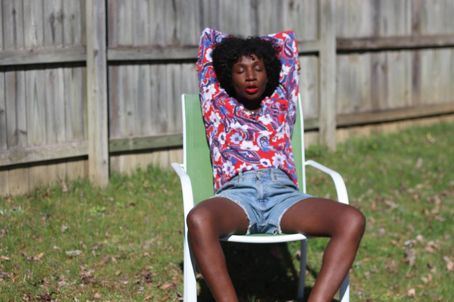 In a bright outdoor setting with a green lawn and a wooden fence, a woman is seated, displaying a vintage long-sleeved blouse by Talbot's. The retro  blouse features a colorful red, white, and blue design and is made from merino wool.