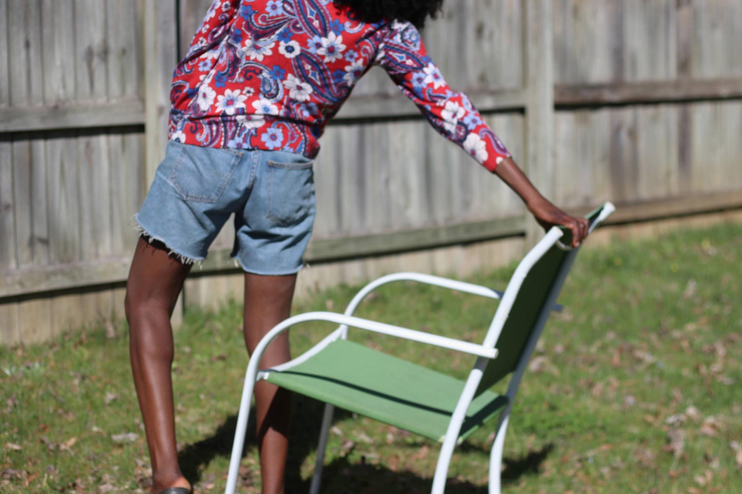 A female model with curly black hair stands next to  a white and green lawn chair on a sunny lawn, showcasing a red, white, and blue patterned vintage Talbot's blouse made of 100% merino wool. A wooden fence and green foliage form the backdrop.
