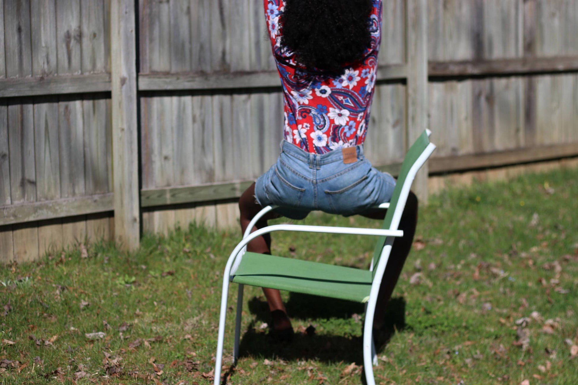 Back view of a woman wearing a vintage Talbot&#39;s merino wool blouse with a distinctive floral pattern is the focal point of a sunny outdoor scene. The setting features a lawn, a chair, and a fence, providing context to the garment.