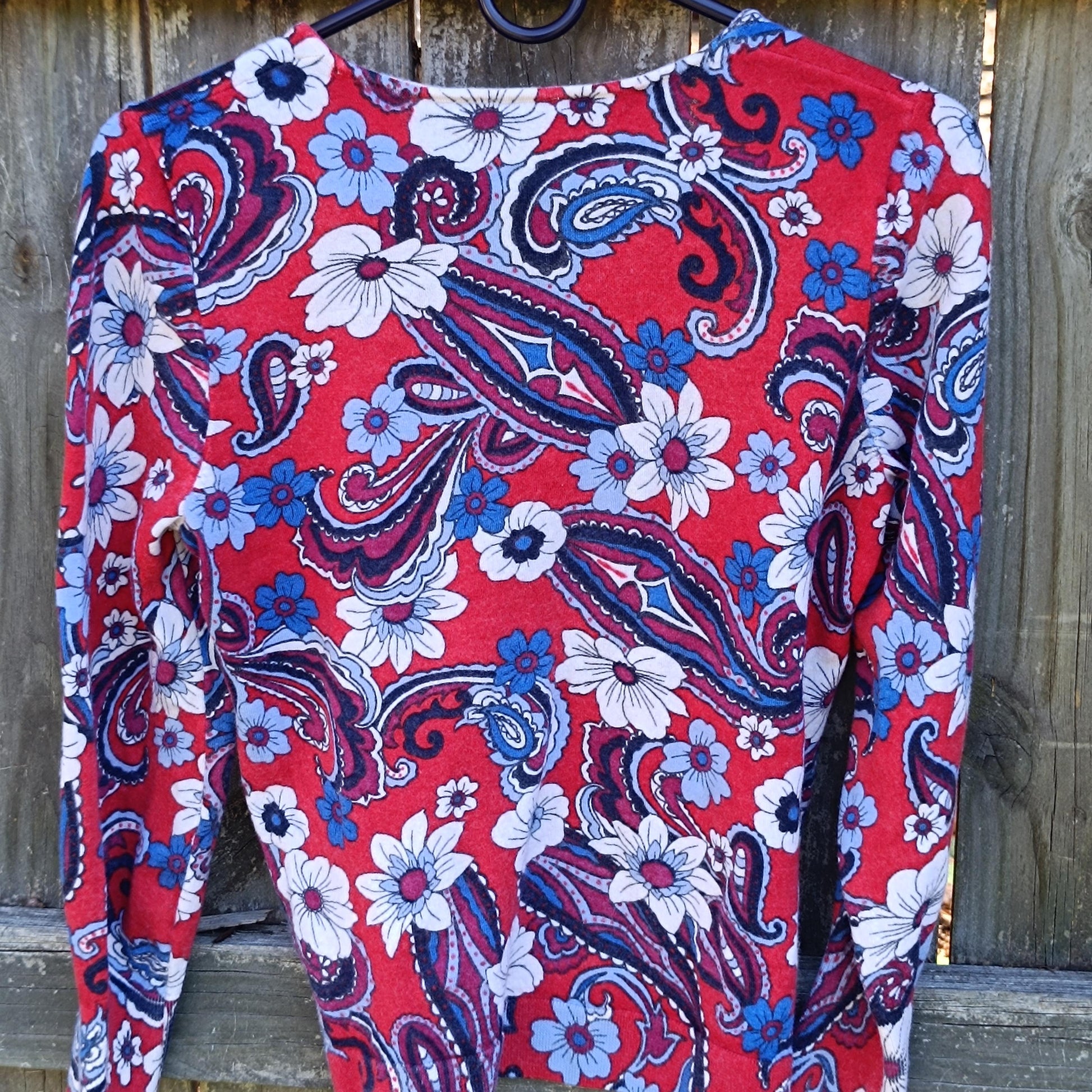 An image of a retro vintage merino wool Talbot&#39;s blouse The red, white, and blue floral top is photographed on a hanger against a brown wooden fence outdoors