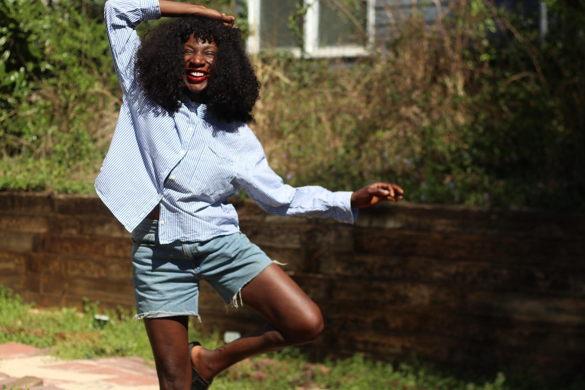 Female model with curly black hair wearing  a striped vintage Old Navy Button up Shirt in blue and white styled with denim cut offs is photographed outdoors against a background with browning grass and a wooden wall infront of a residential white window.