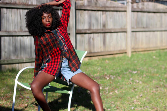 A woman with voluminous curls poses outdoors wearing a vintage Coldwater Creek plaid shirt in red and green. The relaxed-fit button-up, made from a soft viscose-cotton blend, drapes effortlessly over her denim cutoffs, creating a laid-back, edgy look