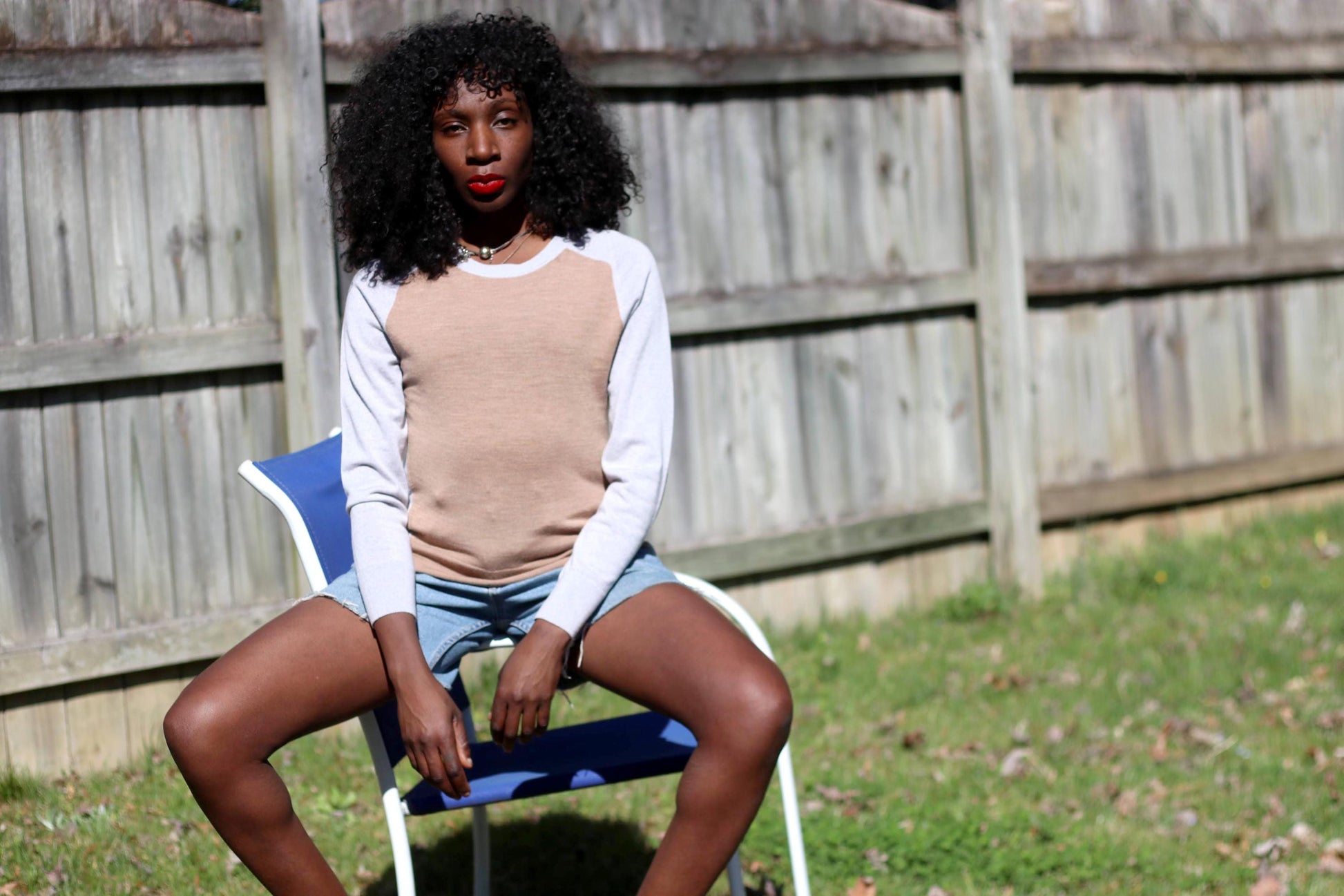 A woman with curly hair sits outdoors on a blue lounge chair in a vintage y2k Banana Republic merino wool sweater, featuring a beige body and gray sleeves. The lightweight knit drapes effortlessly over her frame, styled with denim cutoffs