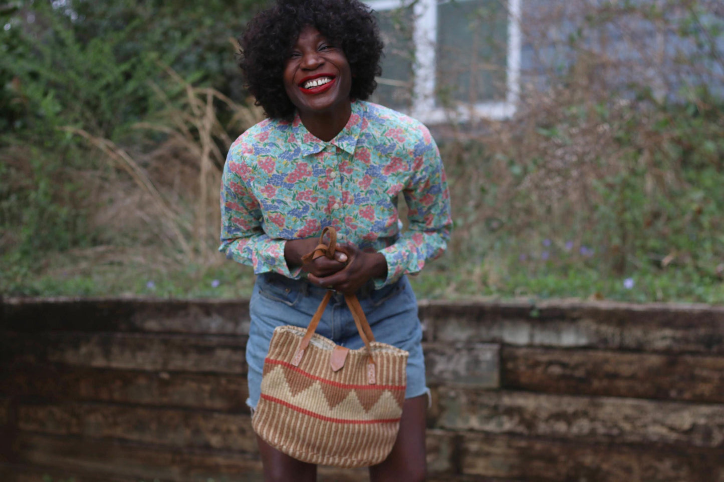 A woman with a radiant smile wears a vintage J.Crew floral button-up shirt, featuring a delicate pink, blue, and green floral print on cotton. She pairs the lightweight collared blouse with high-waisted denim shorts, holding a woven tote bag