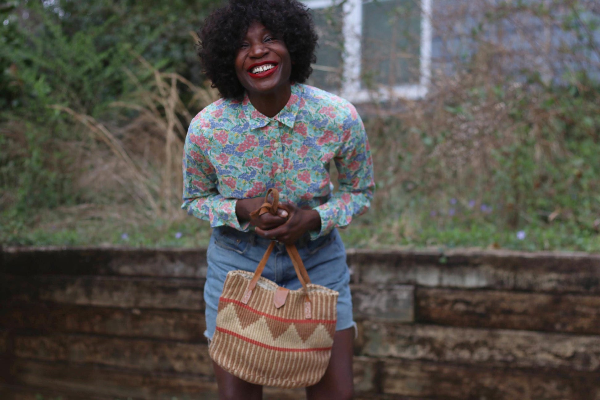 A woman with a radiant smile wears a vintage J.Crew floral button-up shirt, featuring a delicate pink, blue, and green floral print on cotton. She pairs the lightweight collared blouse with high-waisted denim shorts, holding a woven tote bag
