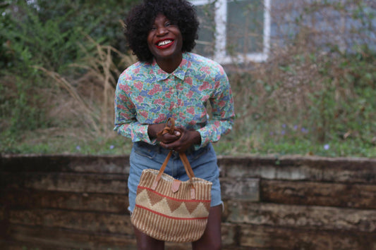 A woman with a radiant smile wears a vintage J.Crew floral button-up shirt, featuring a delicate pink, blue, and green floral print on cotton. She pairs the lightweight collared blouse with high-waisted denim shorts, holding a woven tote bag