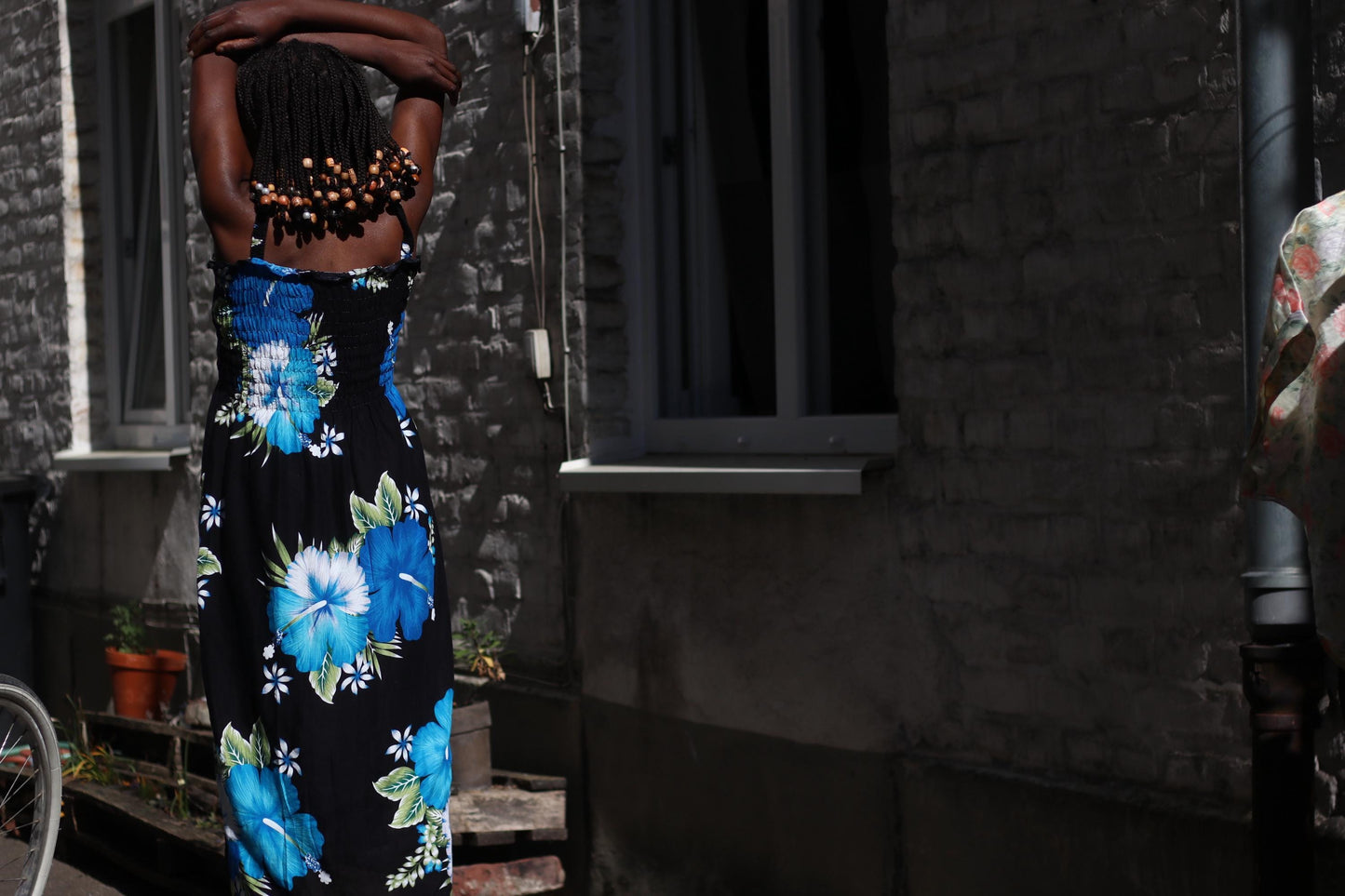 Back view of a sun-drenched image of a Black model wearing a 90s  lightweight rayon dress with rouched bustline, thin straps and a flowing silhouette. The dress features a bold black canvas decorated with large, eye-catching blue and white floral prints. Modem stands in front of a rustic stone building with windows and potted plants lining