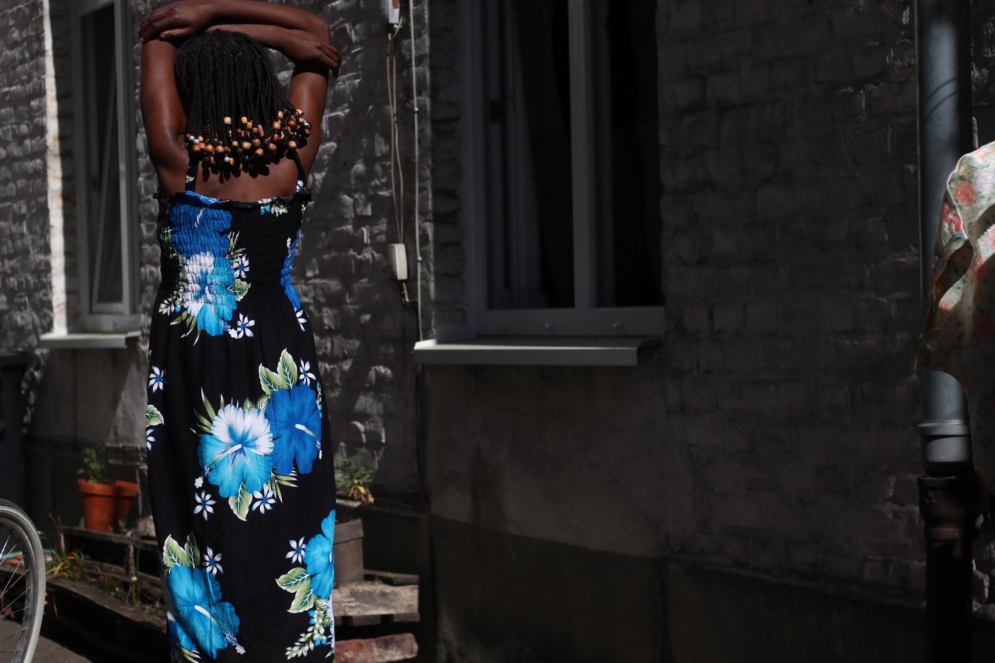 Back view of a sun-drenched image of a Black model wearing a 90s  lightweight rayon dress with rouched bustline, thin straps and a flowing silhouette. The dress features a bold black canvas decorated with large, eye-catching blue and white floral prints. Modem stands in front of a rustic stone building with windows and potted plants lining