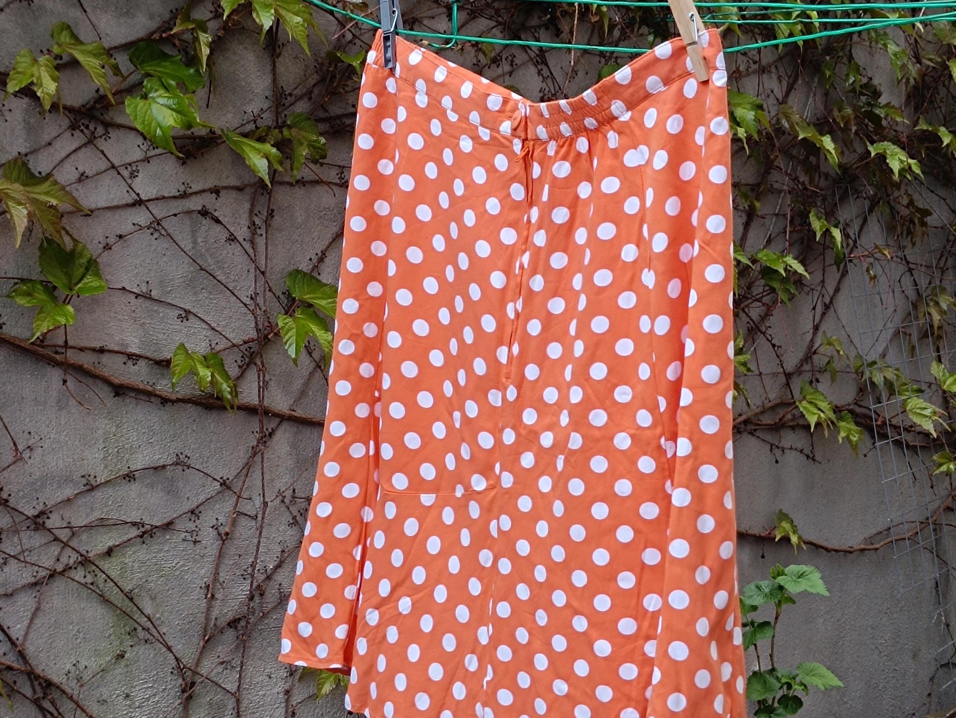 A Chic French Flair 90s Polka Dotted Skirt in Orange is captured on a clothing line outdoors in a garden against a dreamy wall with leavy vines. The  vintage Flirty Mini Skirt excuses a Parisian Summer vibe